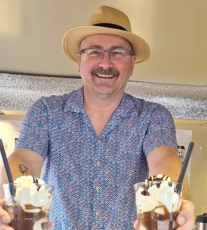 Ein lächelnder Mann mit Strohhut und gemustertem Hemd hält zwei Gläser mit Eisbechern in der Hand, die mit Schlagsahne und Schokoladensirup gefüllt sind.