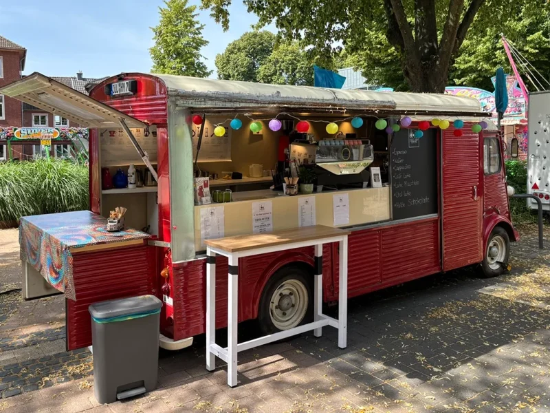 Ein roter Oldtimer-Imbisswagen mit offenem Servierfenster ist mit bunten Lichterketten geschmückt. Draußen gibt es eine Speisekarte, eine Theke, einen Tisch und einen Mülleimer, und die Szene spielt sich in einem schattigen Park ab.