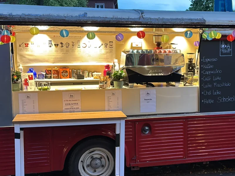 Ein alter roter Imbisswagen mit Serviertheke, bunten Lichterketten und einer Speisekarte auf einer Tafel. Der Truck ist für den Geschäftsbetrieb geöffnet und verfügt über eine Kaffeemaschine und verschiedene Vorräte im Inneren.