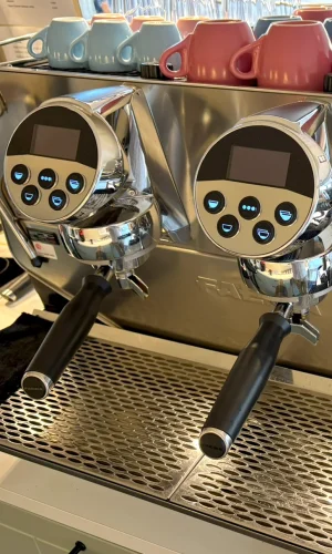 Nahaufnahme einer Espressomaschine aus Edelstahl mit zwei Brühgruppen und digitaler Steuerung. Auf der Maschine sind bunte Tassen gestapelt, vor allem rosa und blau.
